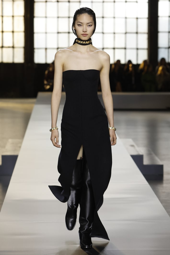 The model wears a dark-grey wool long strapless dress, a black silk ribbon with GG pattern, a choker with gold studs, a bracelet with gold studs, and over-the-knee boots in black leather from the Gucci Women's Fall-Winter 2024 collection.