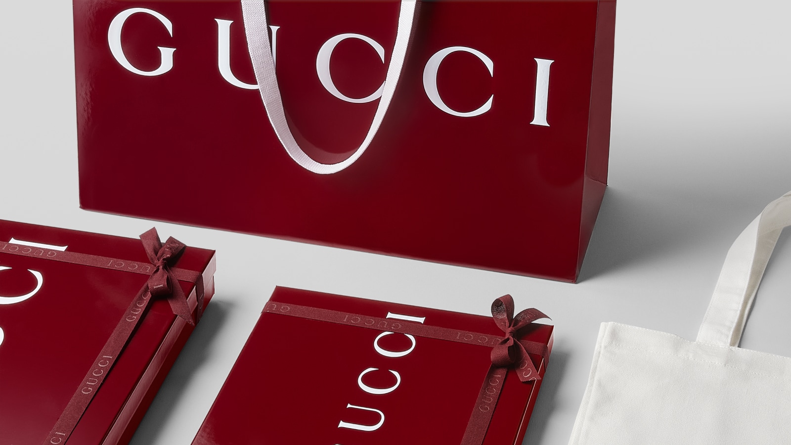 Gucci red shopping bag surrounded by matching different sizes of Gucci signature packaging and a white cotton canvas bag with Gucci logo in white.