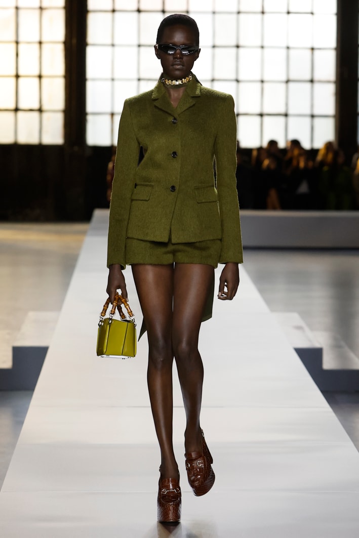 Das Model trägt folgende Designs aus der Gucci Herbst Winter 2024 Damenkollektion: einen olivgrünen Blazer aus Wolle, olivgrüne Shorts aus Wolle, eine Sonnenbrille in Katzenaugenform aus schwarzem Azetat, einen Mini-Shopper aus senfgelbem Leder mit Bambusgriffen, eine Choker-Halskette mit goldfarbenen Nieten und Loafer mit hohem Absatz, Plateausohle und Fersenriemchen aus dunkelbraunem Leder