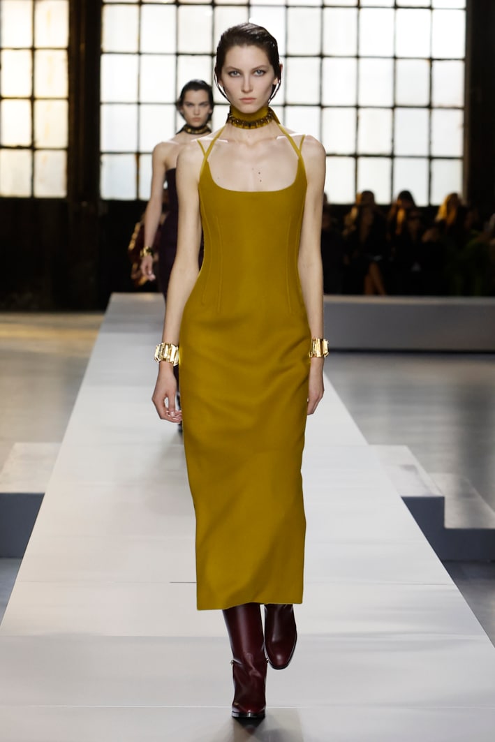 The model wears a mustard wool dress with a slit at the back, a mustard silk ribbon with GG pattern, a choker with gold studs, a bracelet with gold studs, and over-the-knee boots in dark red leather with delicate lightweight gold horsebit detail from the Gucci Women's Fall-Winter 2024 collection