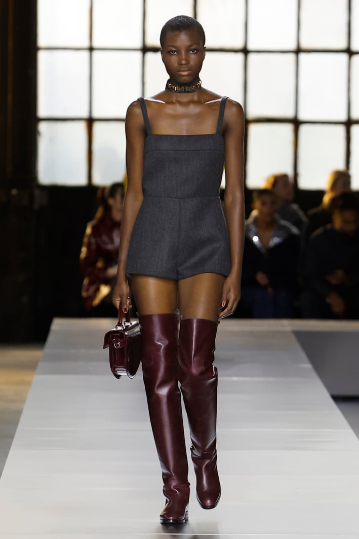 Das Model trägt folgende Designs aus der Gucci Herbst Winter 2024 Damenkollektion: einen grauen Playsuit mit quadratischem Ausschnitt, ein graues Seidenband mit GG Muster, eine große Henkeltasche aus glänzendem Leder in Rosso Ancora mit Doppel G Verschluss und eine Choker-Halskette mit goldfarbenen Nieten