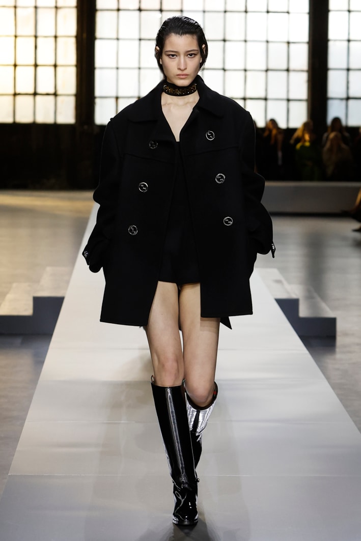 Das Model trägt folgende Designs aus der Gucci Herbst Winter 2024 Damenkollektion: einen schwarzen, übergroßen Dufflecoat aus Wolle, einen schwarzen Playsuit aus Wolle mit quadratischem Ausschnitt, ein schwarzes Seidenband mit GG Muster, eine Choker-Halskette mit goldfarbenen Nieten und vom Reitsport inspirierte Stiefel aus schwarzem, hochglänzendem Leder.