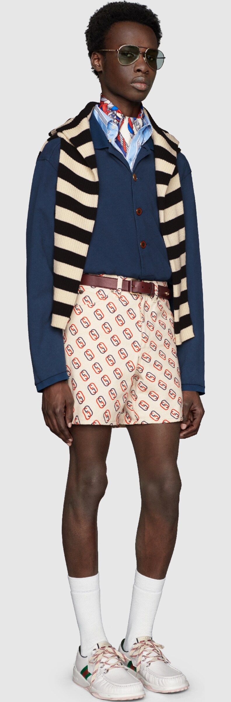 A model wearing Gestreiftes Baumwollhemd mit Bienen-Fil Coupé, Shorts mit ovalem GG Print, Pilotensonnenbrille aus Metall, Tuch aus Seide mit Löwen- und Ketten-Print, Gestreifter Pullover aus Baumwolle mit Lyra, Schnürschuh aus Leder mit Web, Poloshirt aus Baumwoll-Piqué