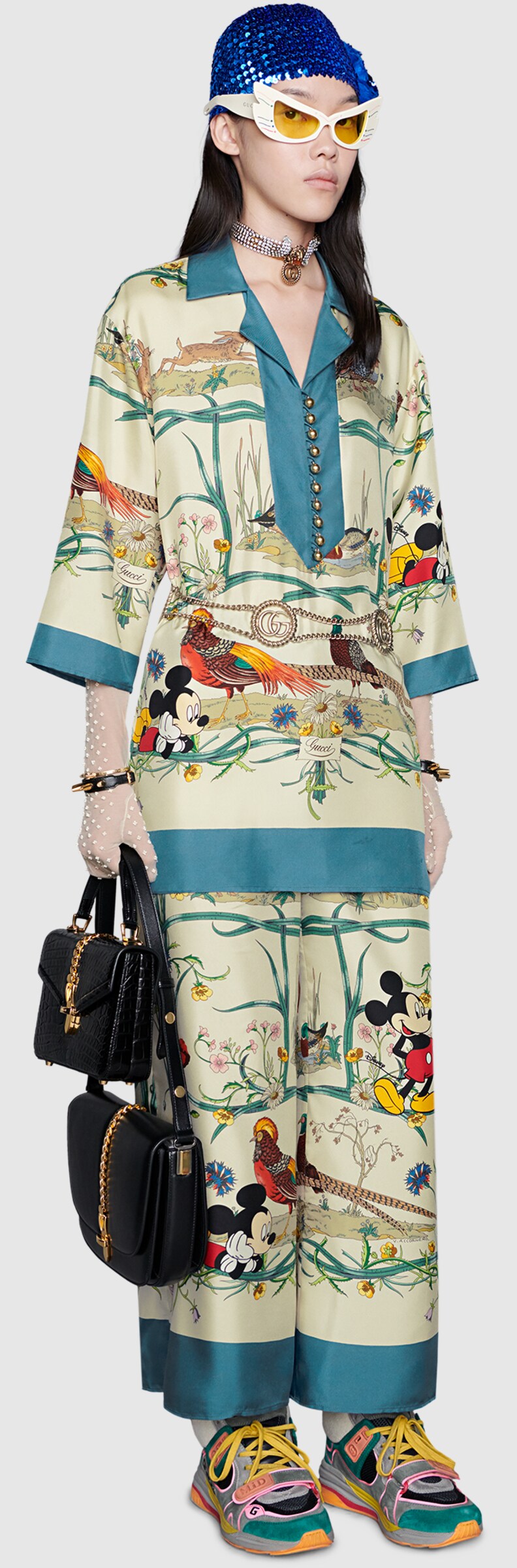 A model wearing Mask acetate sunglasses, Sylvie 1969 small shoulder bag, GG and floral motif lace tights, Chain belt with torchon Double G, Disney x Gucci silk shirt, Disney x Gucci silk pyjama trousers , Women's Ultrapace mid-top sneaker, Lion head choker with Double G
